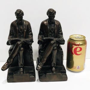 May include: A pair of bronze bookends featuring a seated figure of Abraham Lincoln holding a book. The bookends are detailed and have a realistic appearance.