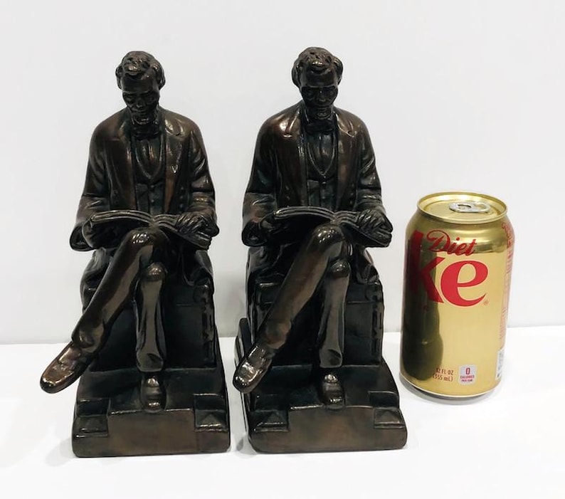 May include: A pair of bronze bookends featuring a seated figure of Abraham Lincoln holding a book. The bookends are detailed and have a realistic appearance.