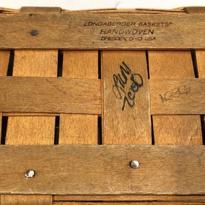 May include: A close-up of a wooden basket with the text "Longaberger Baskets" and "Handwoven Dresden O-O USA" printed on the top. The basket is made of natural wood and has a woven pattern. The text "July 2000" is handwritten on the basket.