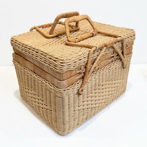 May include: A woven wicker picnic basket with a hinged lid and two handles. The basket is made of natural, light brown wicker and has a square shape.