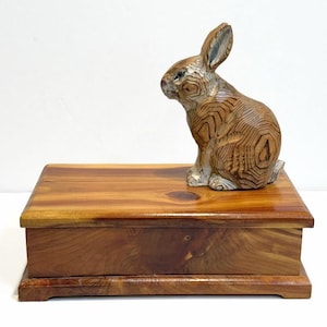 May include: A wooden box with a carved wooden rabbit figurine on top. The box is rectangular with a hinged lid and a dark brown finish. The rabbit is sitting upright with detailed carved patterns.