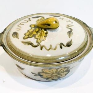 May include: A ceramic serving dish with a lid. The dish is white with a floral design and green and brown accents. The lid features a yellow fruit-shaped handle and leaf design. The dish has two handles on either side.