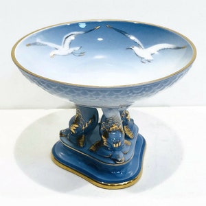 May include: A blue and white porcelain bowl with a scalloped edge and a gold rim. The bowl is decorated with two seagulls in flight and has a base with three fish-shaped legs.