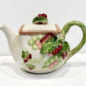 May include: Cream-colored ceramic teapot with a textured basketweave design. The teapot features a lid with a red flower and green leaf detail. A cluster of grapes and leaves adorns the side, with a green vine handle.