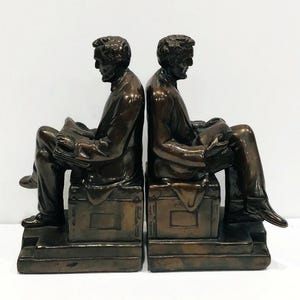 May include: A pair of bronze bookends featuring a seated figure of Abraham Lincoln reading a book. Each bookend is on a base with a rectangular plaque.