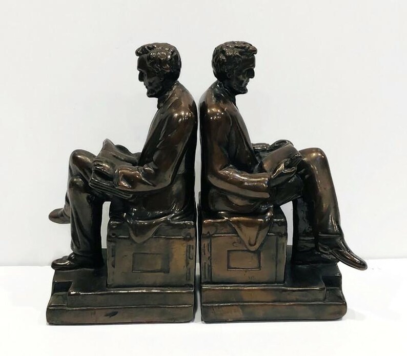 May include: A pair of bronze bookends featuring a seated figure of Abraham Lincoln reading a book. Each bookend is on a base with a rectangular plaque.
