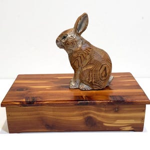 May include: A wooden rabbit figurine with detailed carved patterns sits atop a rectangular wooden box. The rabbit is light brown with darker brown detailing. The box is a rich, reddish-brown color.