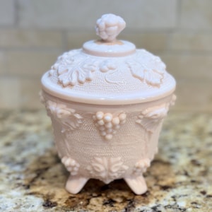 May include: A pink glass candy dish with a grapevine design and three feet. The lid has a grape cluster finial.
