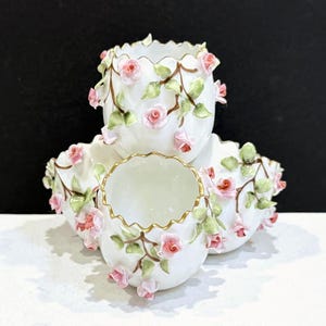 May include: White porcelain decorative bowls with pink rose and green leaf accents. The bowls have a scalloped edge with gold trim. The roses and leaves are arranged around the exterior of the bowls.