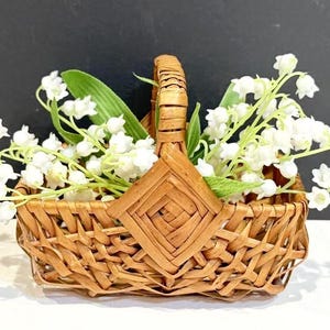 May include: A small, woven wicker basket filled with white lily of the valley flowers and green leaves. The basket has a handle and a diamond-shaped design on the front. The background is a dark gray.