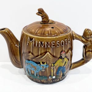 May include: Brown ceramic teapot with a squirrel handle and lid finial. The teapot is decorated with the word "MINNESOTA" and a scene of Paul Bunyan and a blue ox. The base of the teapot reads "PAUL BUNYAN".