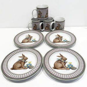 May include: Set of four white ceramic dinner plates with a black and white checkered border. Each plate features a brown bunny holding a blue and yellow Easter egg. The plates are part of a larger Easter themed dinnerware set.