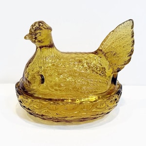 May include: Amber-colored glass hen on a nest-shaped dish. The hen has textured details and a detailed tail. The dish has a decorative, woven-like pattern around the base. A decorative kitchen or dining room accent.