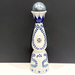 May include: A tall, cream-colored ceramic bottle with a rounded base and a slender neck. The bottle features hand-painted blue leaf patterns and a central floral emblem. It is topped with a smooth, metallic silver, dome-shaped bottle stopper.
