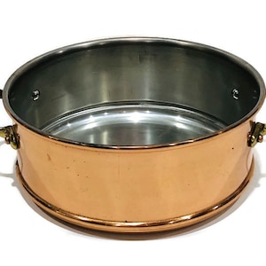 May include: A copper cooking pot with brass handles. The pot has a shiny, metallic finish and is designed for stovetop cooking.