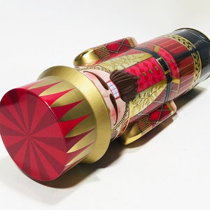 May include: A red and gold nutcracker tin with a festive design. The nutcracker is wearing a red and gold hat and has a white beard. The tin is decorated with a red and gold plaid pattern.