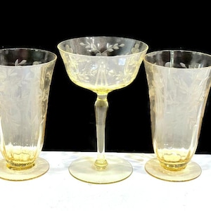 May include: Three vintage yellow-tinted glass drinkware pieces. Includes two tall glasses and a stemmed champagne coupe. All pieces feature etched floral designs. The glasses have a flared shape and round bases.