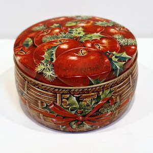 May include: A round tin container with a red, green, and gold design. The lid features a basket of red apples with green holly leaves. The container is perfect for storing small items.