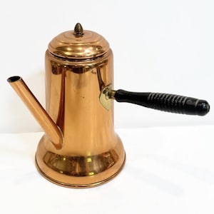 May include: A copper coffee pot with a black wooden handle and a spout. The pot has a lid with a small knob on top. The copper has a shiny finish. The handle is attached with brass hardware.