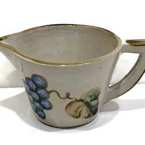 May include: A white ceramic creamer with a brown handle and a grape and leaf design. The creamer is decorated with blue, brown, and green paint.