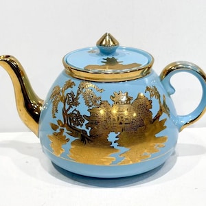 May include: A light blue teapot with gold accents. The teapot features a gold-colored spout, handle, and lid trim. The body of the teapot is decorated with a gold Asian-inspired landscape design. The lid has a gold finial.