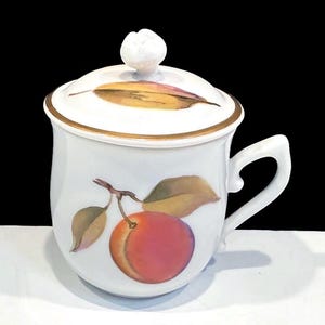May include: White ceramic teacup with a lid, featuring a hand-painted design of a peach and leaves. The lid has a gold trim and a flower-shaped handle. The teacup has a curved handle and a gold trim around the rim.