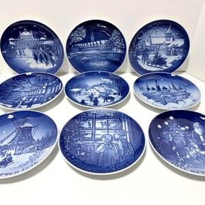May include: A collection of nine circular porcelain plates featuring blue and white winter scenes. Each plate displays a unique detailed illustration of snowy landscapes, buildings, or holiday figures, with some including text like JULAFTEN.
