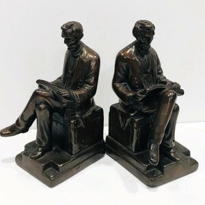 May include: A pair of bronze bookends featuring two seated figures reading books. The figures are dressed in suits and have a realistic appearance. The bookends are a decorative and functional addition to any bookshelf.