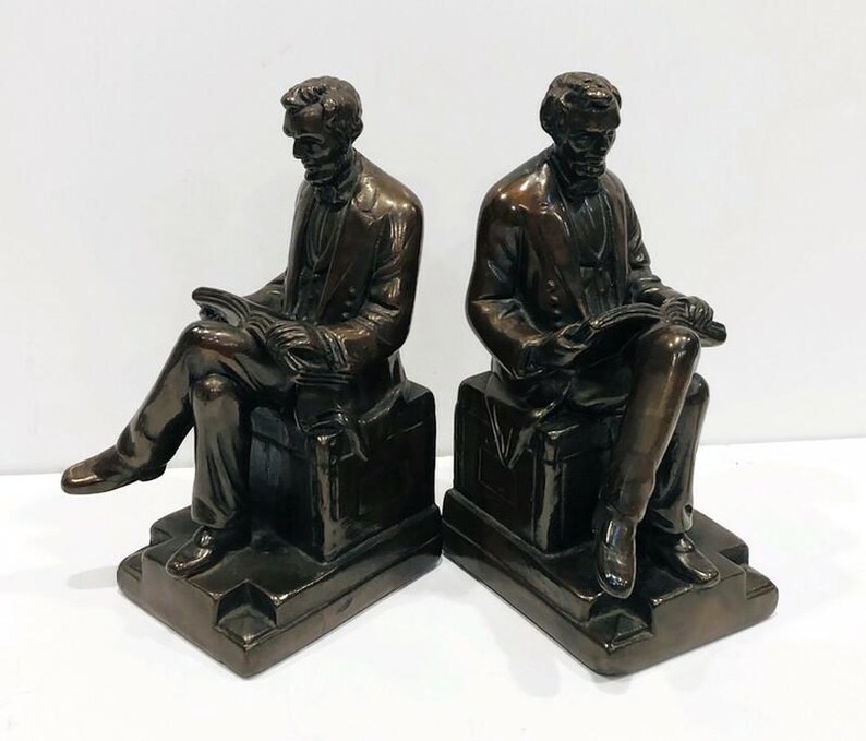 May include: A pair of bronze bookends featuring two seated figures reading books. The figures are dressed in suits and have a realistic appearance. The bookends are a decorative and functional addition to any bookshelf.