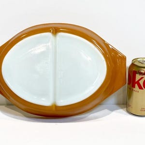 May include: An oval, divided glass serving dish with a glossy butterscotch orange exterior and a white interior, featuring integrated side handles. A 12 fluid ounce aluminum soda can is positioned next to the dish for a visual size reference.