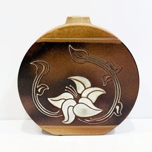 May include: A ceramic vase with a flat, round body and a short neck. The vase is primarily brown with a white floral design. The flower is in the center, with a stem and leaves that curve around it. The vase is approximately 8 inches tall.