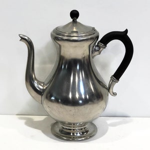 May include: A silver teapot with a black handle and a black knob on the lid. The teapot has a rounded body and a flared spout.