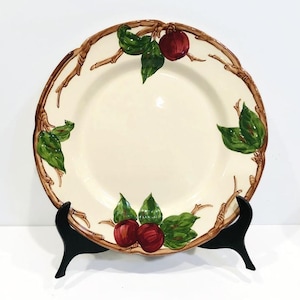 May include: A white ceramic dinner plate with a brown twig and green leaf design. The plate features red apples with green leaves.