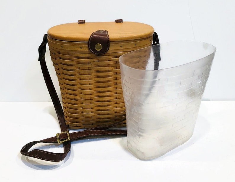 May include: A brown wicker basket with a leather strap and a clear plastic liner. The basket has a lid and is designed to be carried on the shoulder.