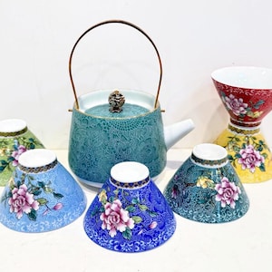 May include: A teal ceramic teapot with a bronze handle and lid, surrounded by six decorative tea cups. The cups are in various colors, including green, blue, and yellow, and feature floral designs. The tea set is likely for serving tea.