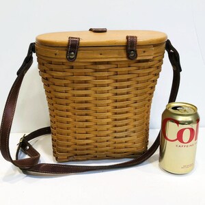 May include: A brown wicker basket with a leather strap and a wooden lid. The basket is about 12 inches tall and 8 inches wide. The basket is sitting on a white surface.