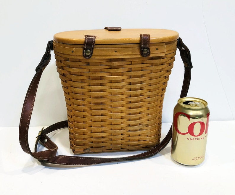 May include: A brown wicker basket with a leather strap and a wooden lid. The basket is about 12 inches tall and 8 inches wide. The basket is sitting on a white surface.