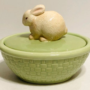 May include: A light green ceramic basket-shaped container with a lid, topped with a small, cream-colored bunny figurine. The basket has a woven texture, and the lid is a solid light green. The bunny has detailed features.