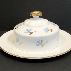 May include: White ceramic butter dish with a lid featuring a gold knob and blue floral design. The dish has a gold rim.