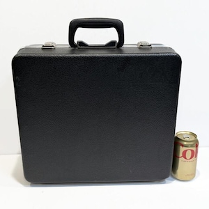 May include: A black, rectangular briefcase with a textured surface, a black handle, and silver-colored clasps. A gold-colored can of soda is next to the briefcase.