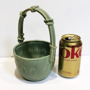 May include: A sage green ceramic basket with a handle, next to a Diet Coke can. The basket has a round bowl shape with a handle that arches over the top. The can is gold with red text that reads "Diet Coke Caffeine Free".