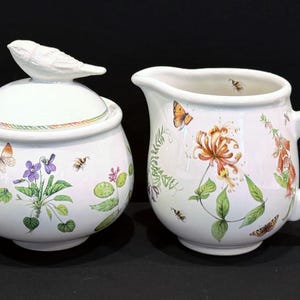 May include: A white ceramic sugar bowl with a bird lid and a matching creamer. Both pieces feature a floral design with butterflies, bees, and green leaves.