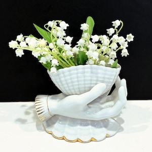May include: A white ceramic vase in the shape of hands holding a shell-shaped bowl filled with white flowers and green leaves. The vase has gold accents on the base and wrist.
