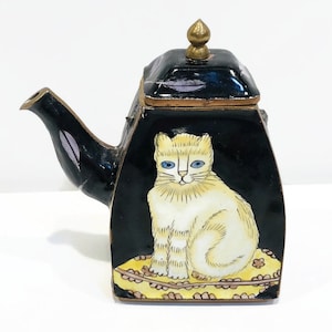 May include: Black teapot with a white cat illustration on the front. The cat has blue eyes and sits on a yellow cushion. The teapot has a gold finial and spout, and a black lid.