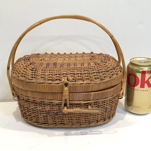 May include: A woven wicker basket with a hinged lid and a handle. The basket has a brown and tan color with red and green accents. The basket is sitting on a white surface next to a can of soda.