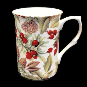 May include: White ceramic mug with a floral pattern featuring red berries, green leaves, and brown pine cones.