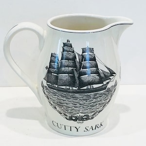 May include: White ceramic pitcher with a black and white illustration of a ship called "Cutty Sark" sailing on the ocean. The ship has three masts and sails. The pitcher has a handle and a spout. The text "Cutty Sark" and "1869" are printed on the bottom of the pitcher.
