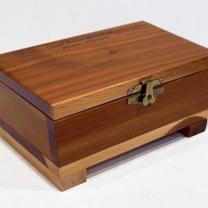 May include: A wooden box with a hinged lid and a brass latch. The box is made of cedar wood and has a natural wood finish. The box is decorated with a simple design and has a small inscription on the top that reads "Crest Saddle Blanket London, Minnesota".