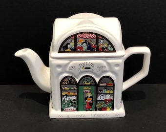 May include: A white ceramic teapot shaped like a cafe, featuring a spout, handle, and lid. The teapot has colorful illustrations of people, food, and the text "Polly's Home Made Teas Sweets and Cakes". The base reads "a cafe teapot".