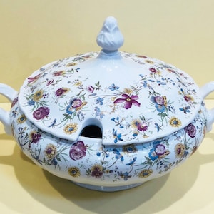May include: White ceramic soup tureen with a lid, adorned with a floral pattern in purple, yellow, and blue. The tureen has two handles and a lid with a finial. A serving spoon slot is visible.